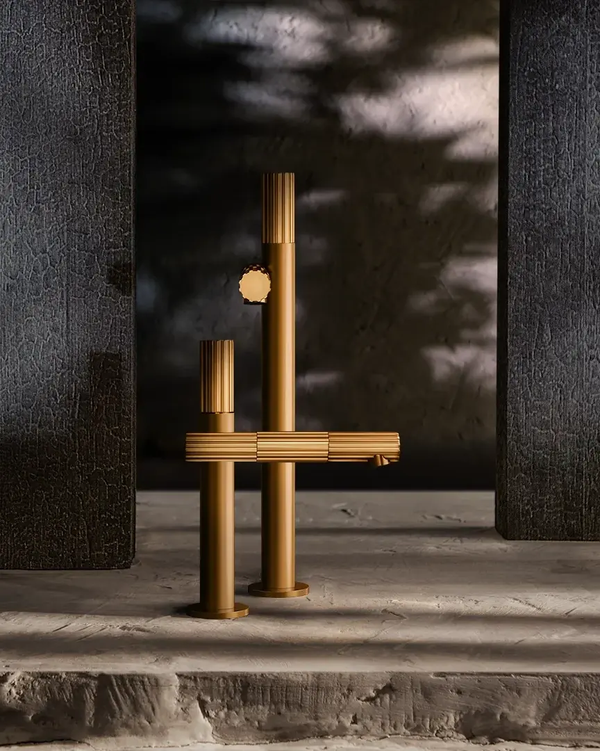 Grifería Gessi Incastri en bronce sobre un suelo de piedra, entre paneles negros texturizados y sombras de hojas